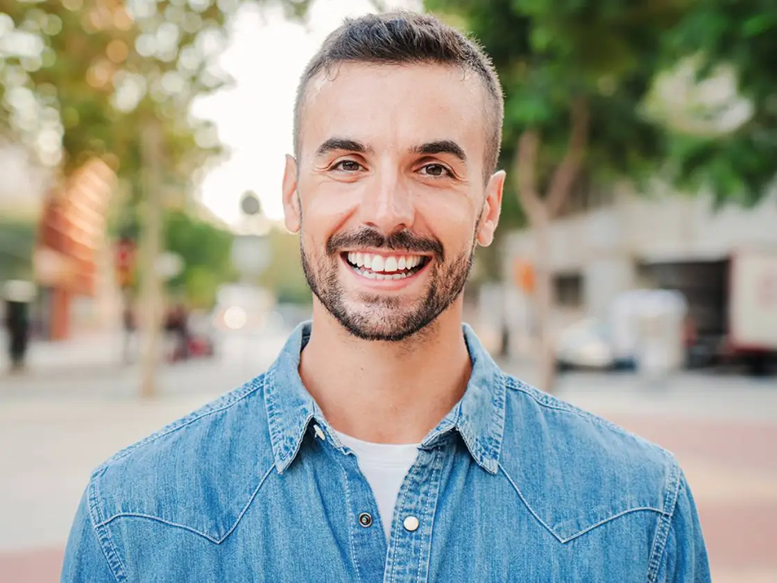 A man with a full, confident smile standing outdoors on a tree-lined street