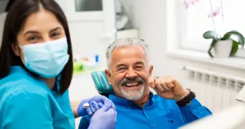 A smiling older patient giving a thumbs up in the dental chair beside a masked dental assistant