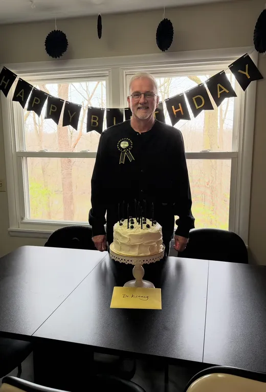 Dr. Kinney behind a birthday cake with black candles under a black and gold Happy Birthday banner